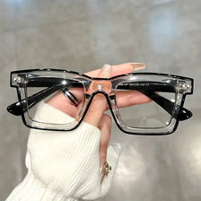 A hand holds a pair of clear square-framed glasses with black arms against a blurred background.