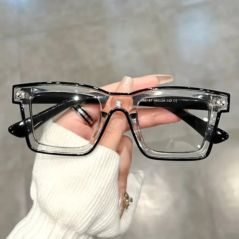 A hand holds a pair of clear square-framed glasses with black arms against a blurred background.