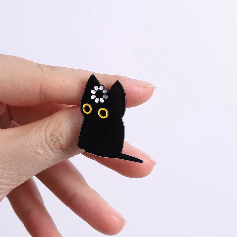 A black enamel pin shaped like a cat with yellow eyes and white dots, held by a hand.