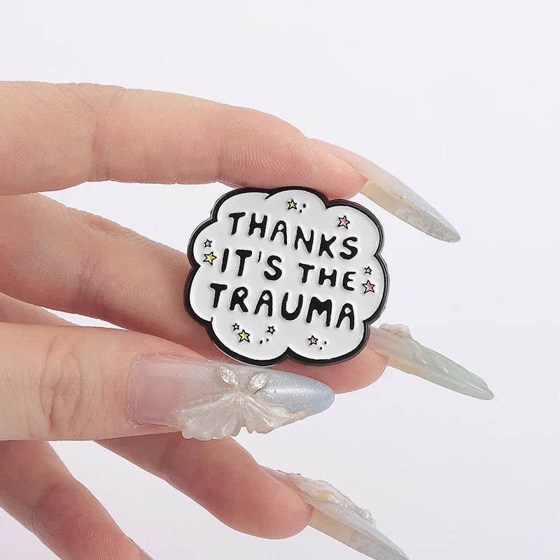 A hand holds a black enamel lapel pin shaped like a cloud with the text Thanks It's the Trauma and small stars.
