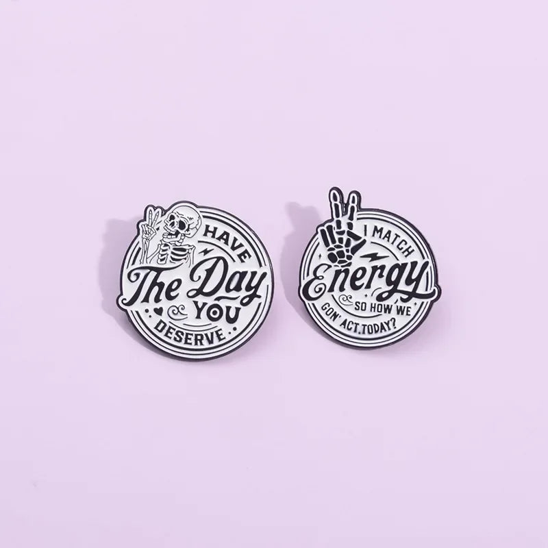 A silver enamel lapel brooch featuring a skull and hand design with the text Have The Day You Deserve and Match Energy So How