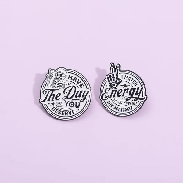 A silver enamel lapel brooch featuring a skull and hand design with the text Have The Day You Deserve and Match Energy So How