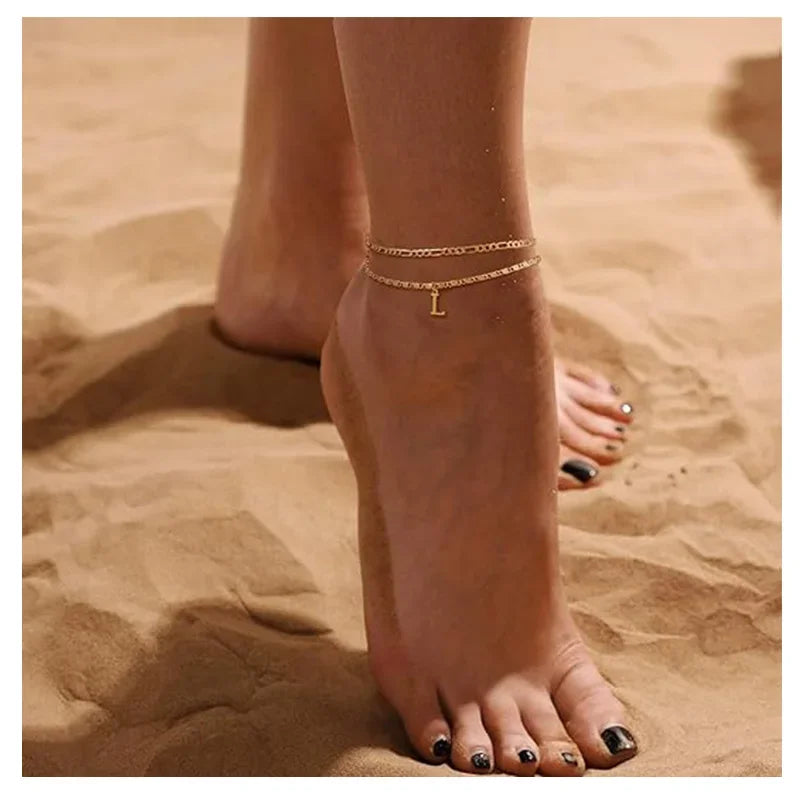 Close-up of a woman's ankle showing layered letter anklets on her foot against a sandy background.