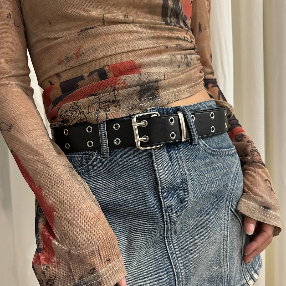 A close-up shot of a person wearing a black leather belt with a silver buckle over denim jeans and a sheer, distressed top.