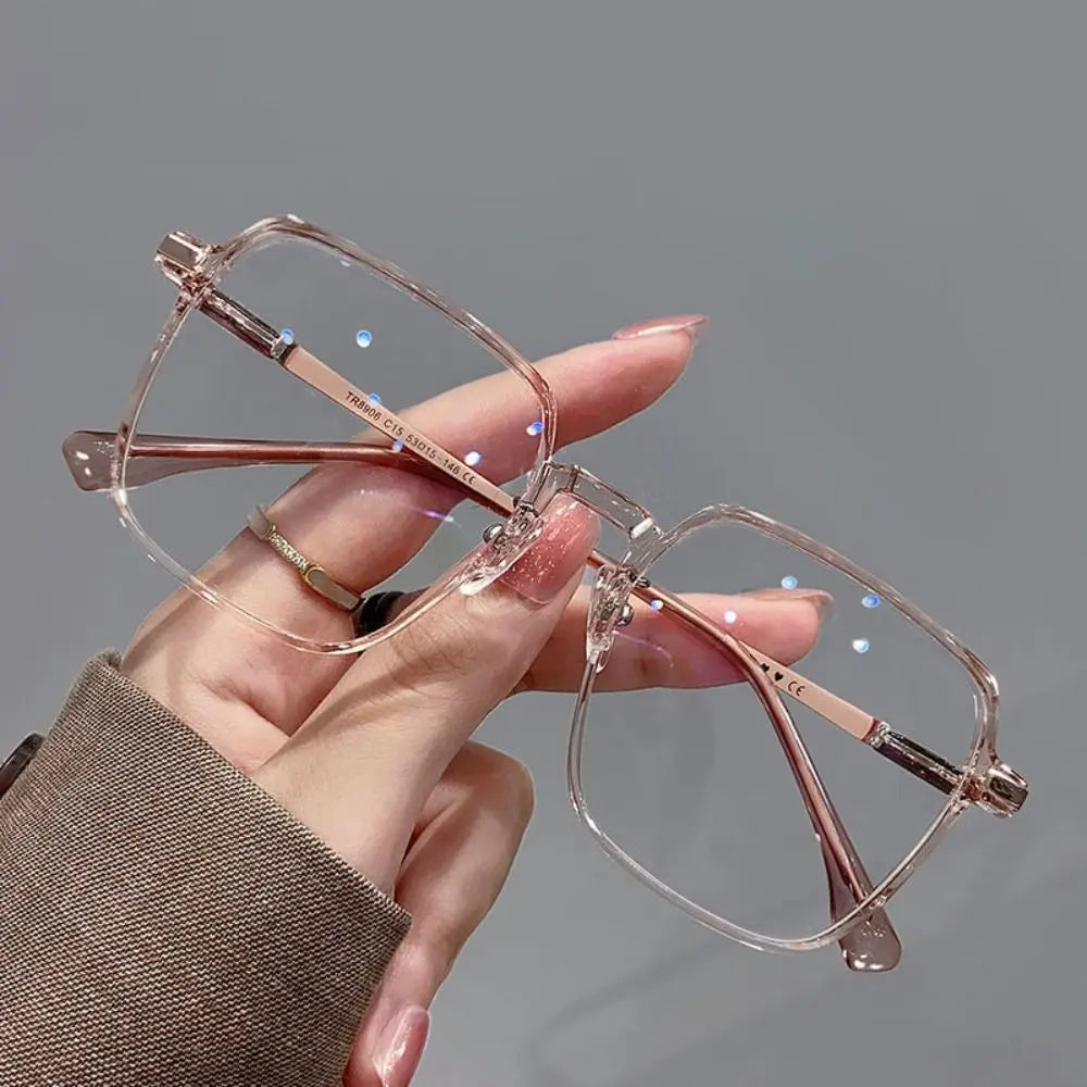 A hand holds a pair of clear metal anti-blue light eyeglasses with rose gold accents against a gray background.