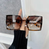 A hand holds a pair of tortoiseshell leopard print square sunglasses against a white background.