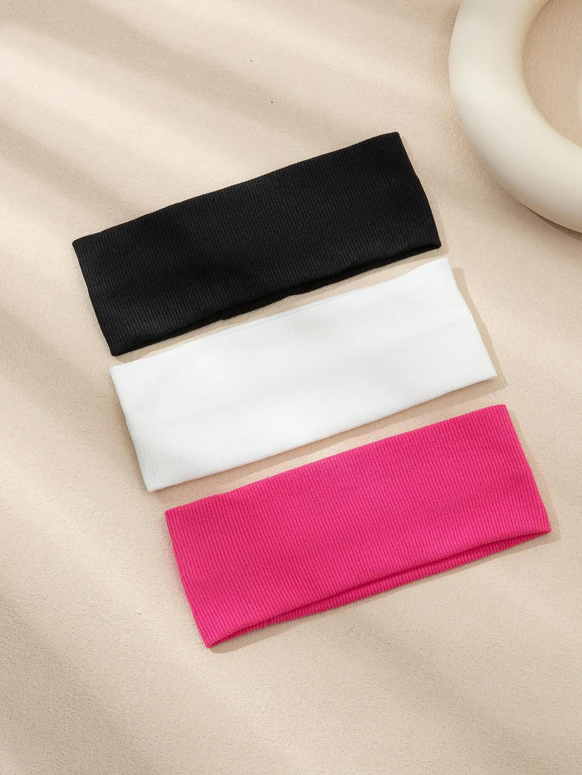A pack of three solid color fashion headbands in black, white, and pink laid on a beige surface.