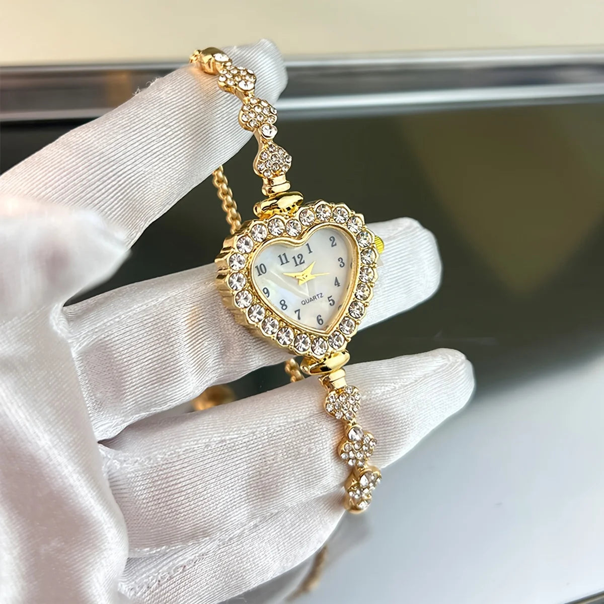 A hand wearing a white glove holds a gold-toned sparkling heart rhinestone quartz watch.