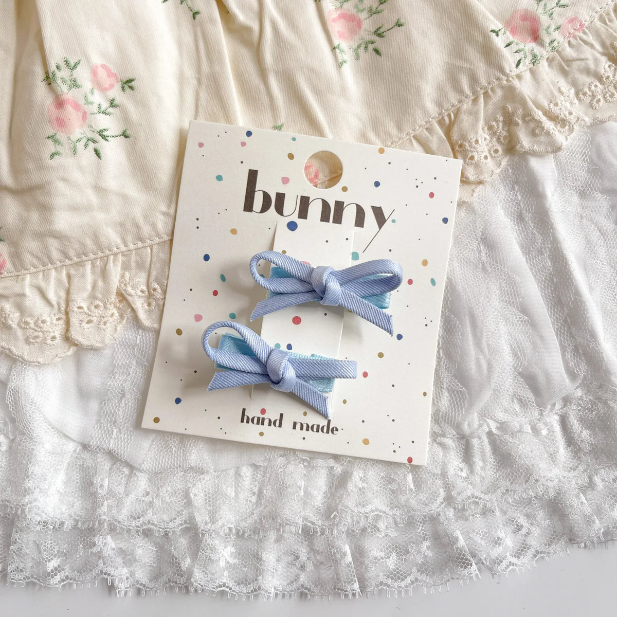 Un ensemble de pinces à cheveux en nœud bleu clair, présenté sur une carte avec un fond de tissu en dentelle blanche.