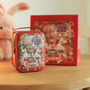 A red tinplate candy box with a carrying loop sits in front of a larger red box featuring a festive holiday scene.