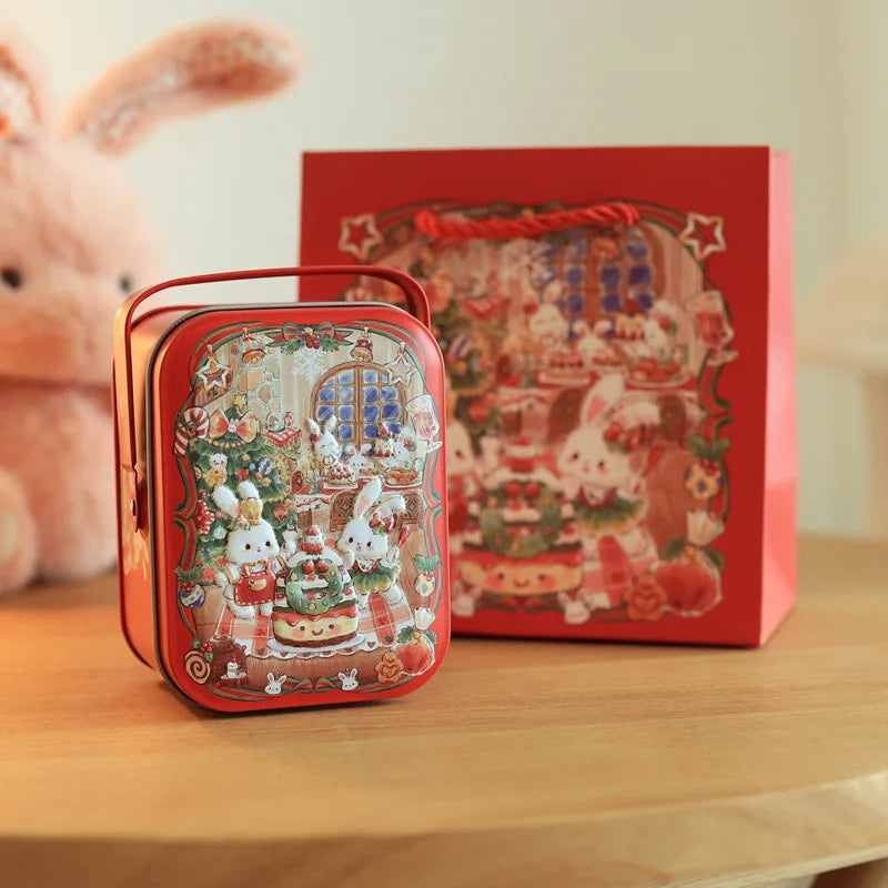 A red tinplate candy box with a carrying loop sits in front of a larger red box featuring a festive holiday scene.