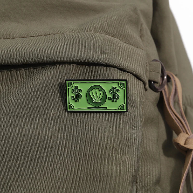 A green enamel pin featuring a custom symbol of US dollars and a leaf design attached to a canvas military-style fabric.