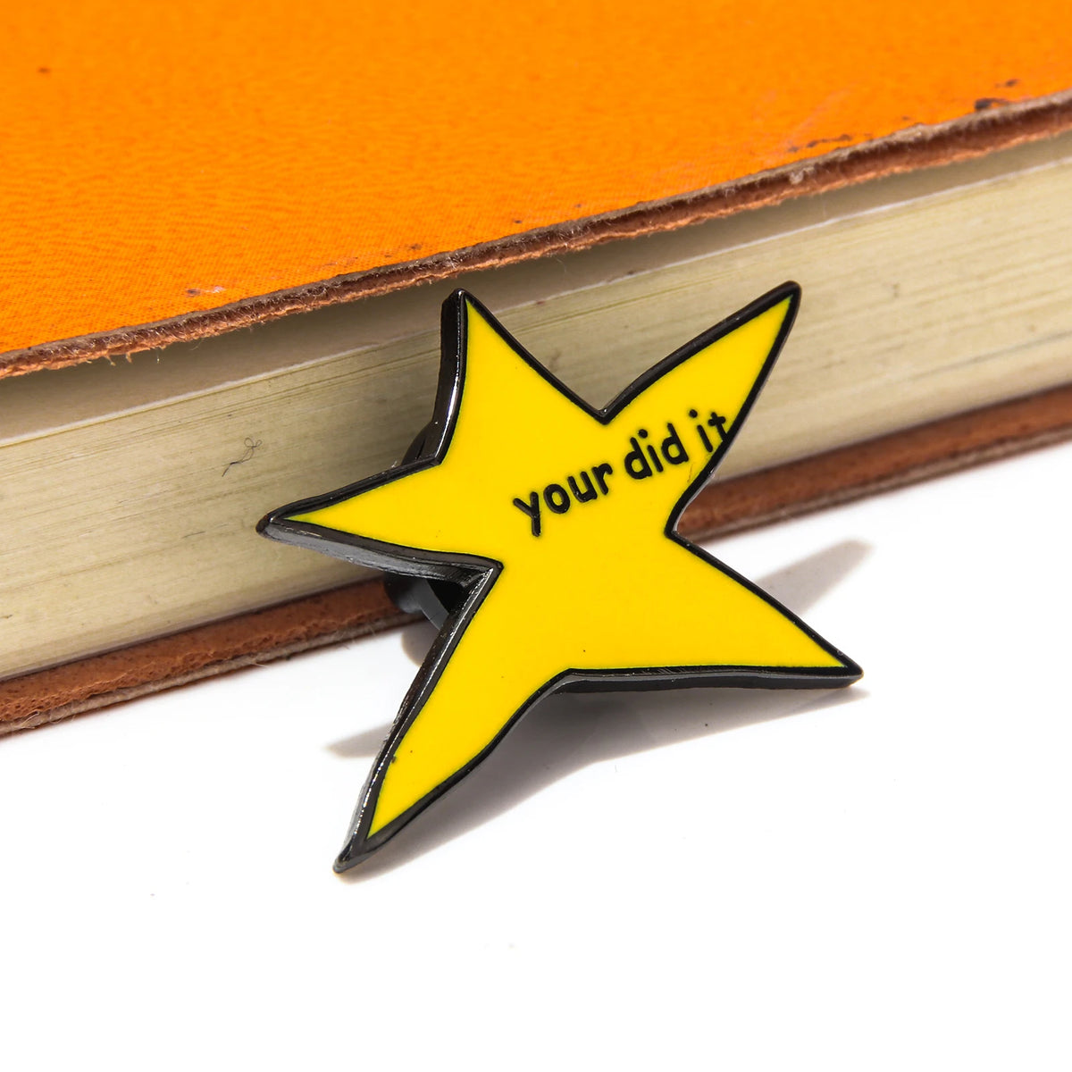 A yellow star enamel pin with the text your did it on a leather-bound book.