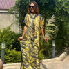 Eine Frau in einem gelb-schwarz gemusterten Boho-Seidenkaftan steht draußen bei grünem Laub.