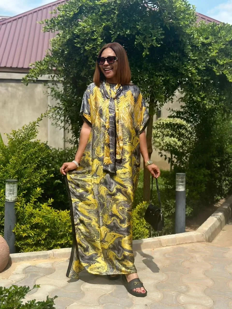Eine Frau in einem gelb-schwarz gemusterten Boho-Seidenkaftan steht draußen bei grünem Laub.
