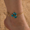 Gold-toned bohemian flower pendant anklet worn on a person's ankle against a sandy background.
