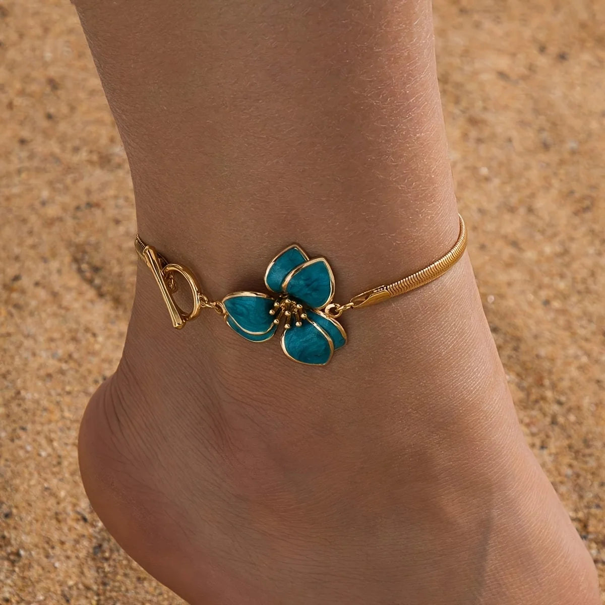 Gold-toned bohemian flower pendant anklet worn on a person's ankle against a sandy background.