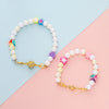 Weißes Perlenarmband mit farbigen Stern-Charms und goldenen Verschluss auf rosa-blauer Fläche.