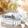 A silver luxury quartz couple watch is displayed on a light surface with soft floral background elements.