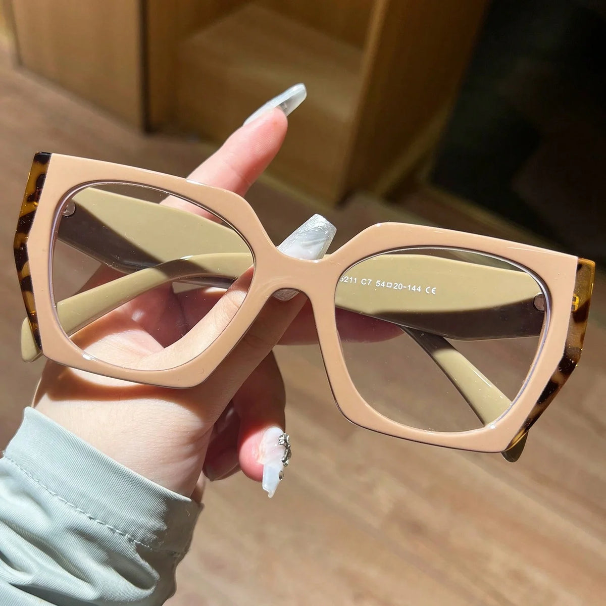 A hand holds a pair of light beige leopard print fashion glasses with large frames.