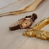 Reloj de correa de cuero marrón retro con decoración dorada y pedrería sobre tela clara junto a un objeto decorativo dorado