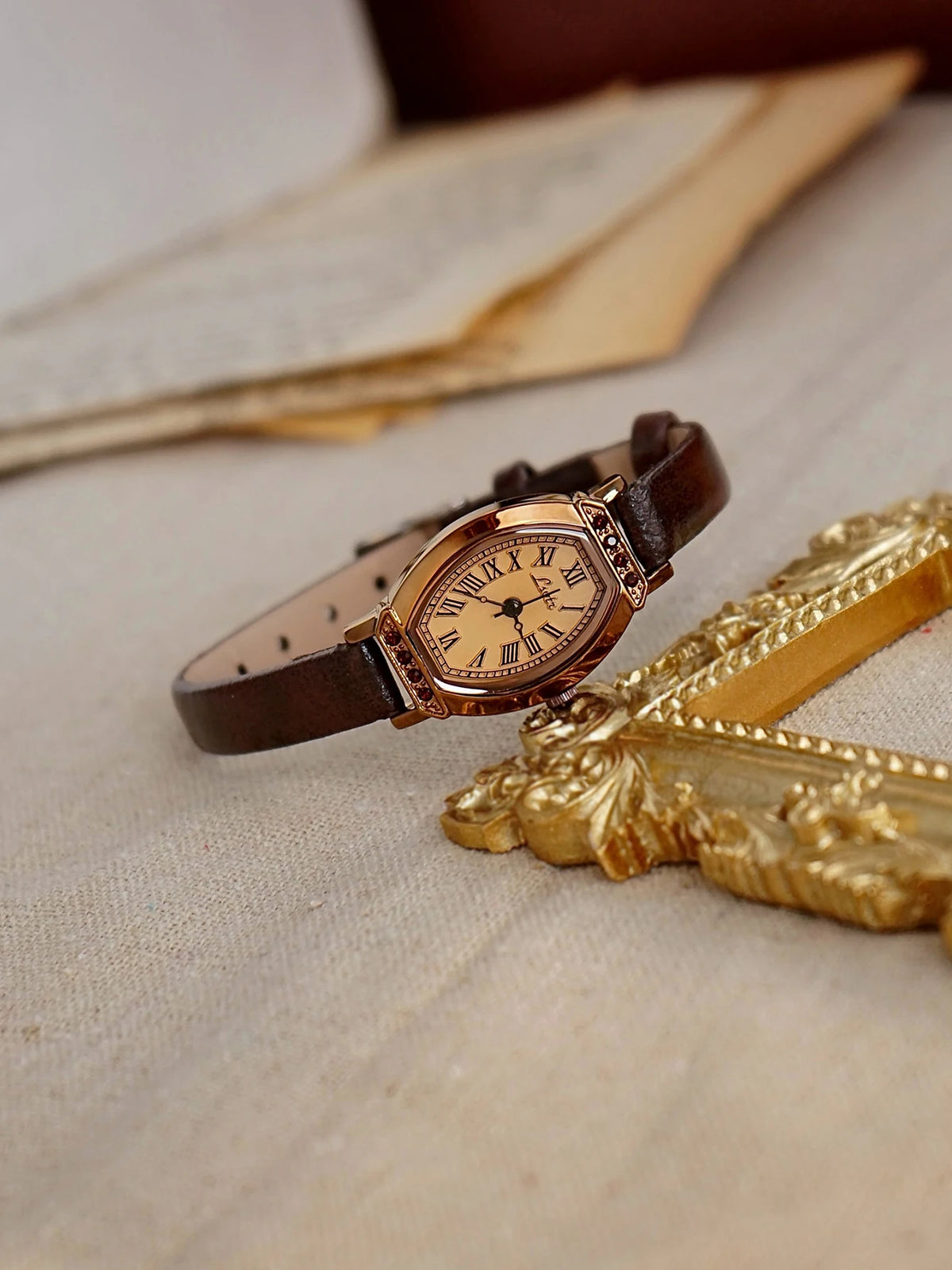 Reloj de correa de cuero marrón retro con decoración dorada y pedrería sobre tela clara junto a un objeto decorativo dorado