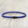 A close-up of a blue thread charm bracelet with a silver clasp resting on a light wooden surface.