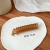 A long, amber-colored rubber braided ponytail holder measuring about 7cm resting on a white ceramic plate.