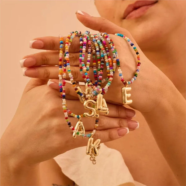 A hand holds a beaded pendant necklace featuring initial letters made of colorful beads.