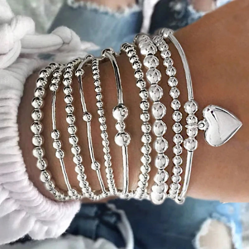 A stack of silver round beaded bracelets worn on a wrist, with a silver heart charm visible.