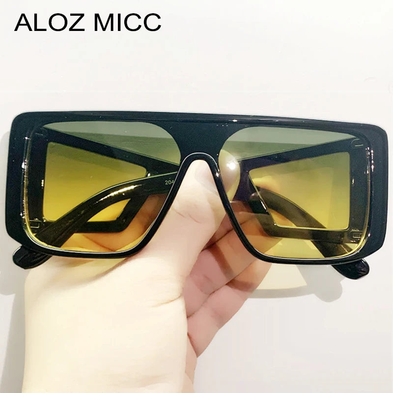 A hand holds a pair of black oversized gradient square sunglasses with yellow-green lenses.