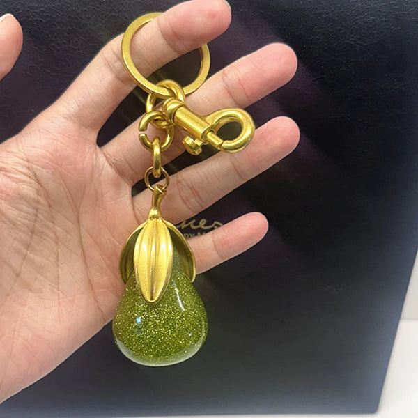 Un charm en forme de poire, ton or, avec une finition pailletée verte, est tenu dans la main d'une personne.