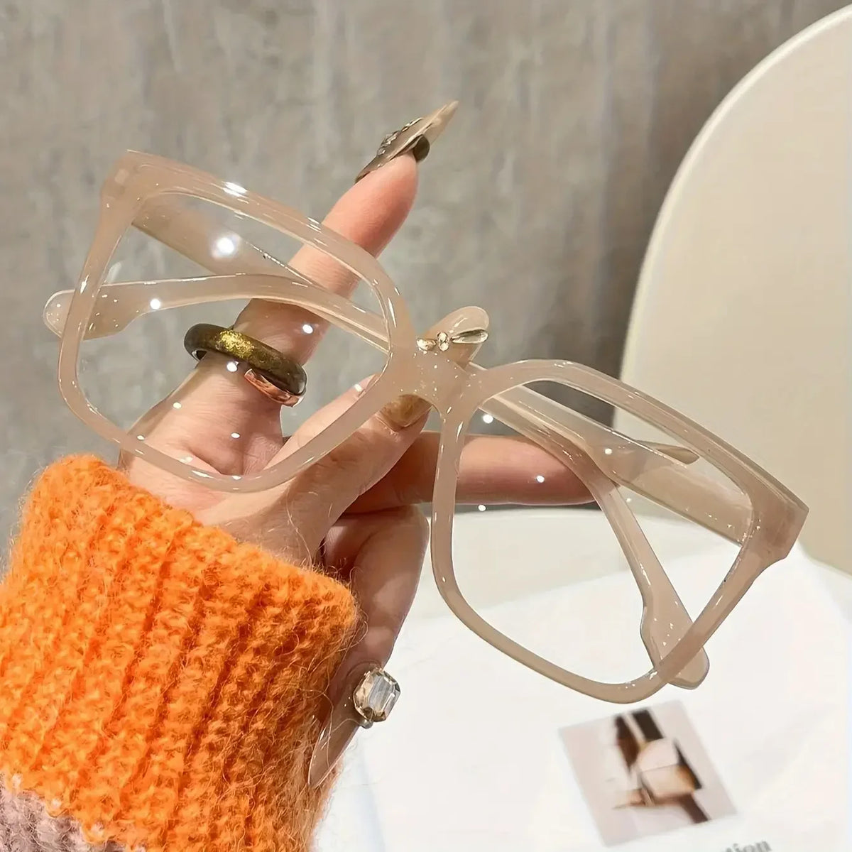 A hand holds a pair of elegant geometric reading glasses with transparent lenses against a blurred background.