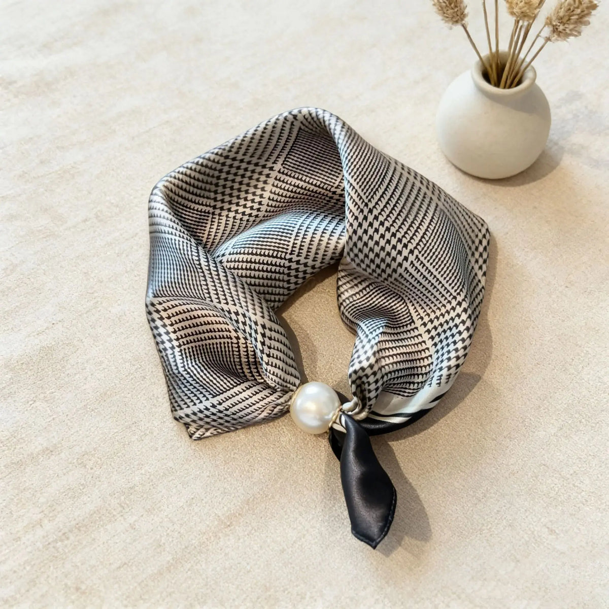 A black and white patterned silk scarf tied with a black ribbon and a pearl detail, resting on a light beige surface.