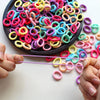 A hand is holding a thin purple elastic band, with many colorful silicone hair bands scattered on a dark plate.