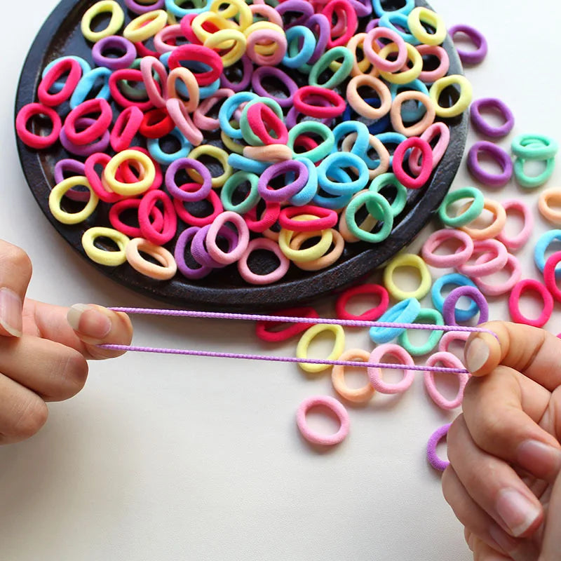 A hand is holding a thin purple elastic band, with many colorful silicone hair bands scattered on a dark plate.