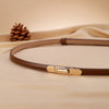 A close-up of a tan skinny leather belt with a gold buckle resting on a beige fabric.