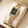 Reloj de pulsera analógico de cuarzo tono dorado con esfera negra y pulsera dorada, exhibido en una caja de color claro.