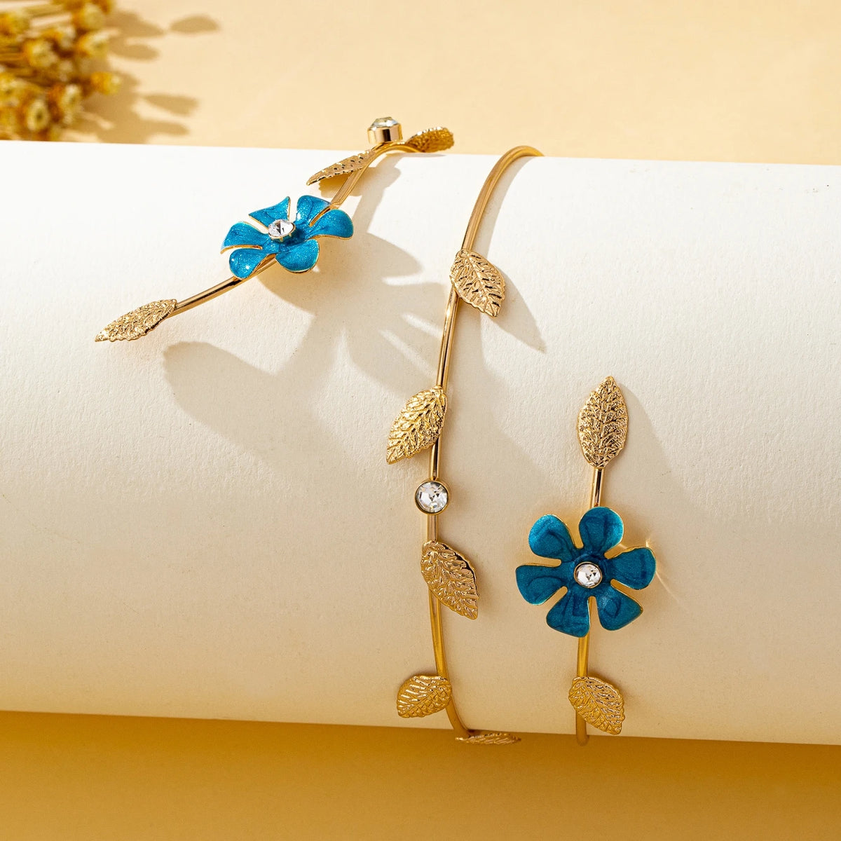 Gold floral vine arm bracelet featuring a blue flower charm and gold leaves on a cream background.