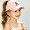 A smiling young girl wearing a pink unicorn baseball cap with a purple design and a light-colored top.