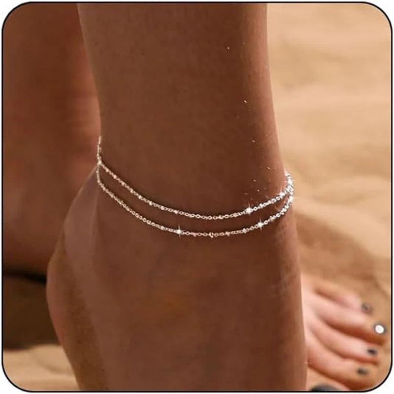 Layered gold and silver plated anklets worn on a foot against a sandy background.