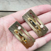 Ein Paar goldfarbener, verschiebbare Mood-Pins mit spitzen, strahlenden Linien wird in der Hand einer Person gehalten.