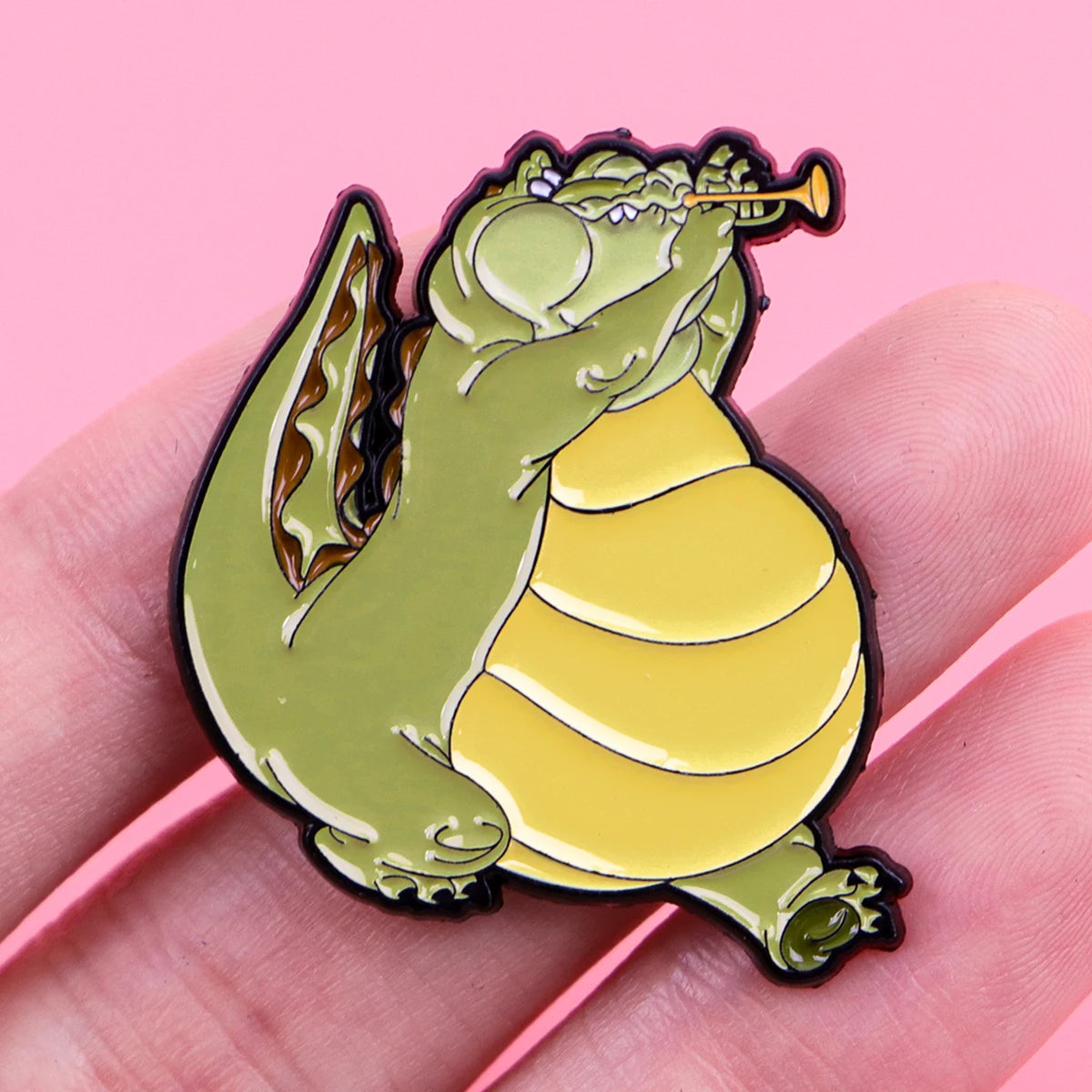 A cute green frog enamel pin with wings is shown being held by a hand.