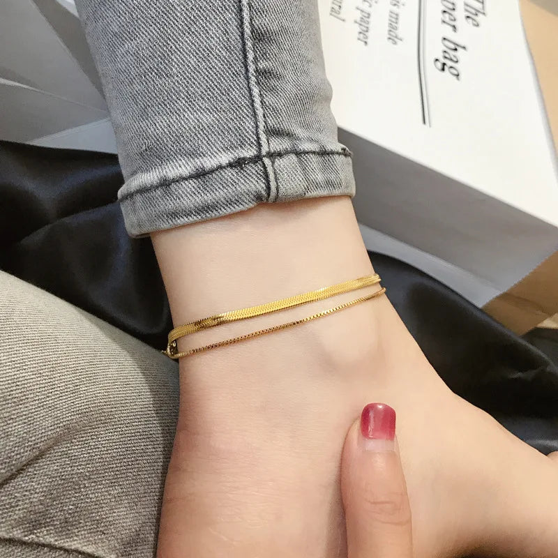 A close-up of a gold double snake anklet worn on a person's ankle, resting near folded gray jeans and a white box.