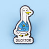 An enamel pin depicting a white duck doctor with a blue uniform and yellow accents.
