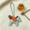 A light blue horse pendant charm with an orange mane is displayed on a cream knitted fabric next to a honey jar and a wooden 