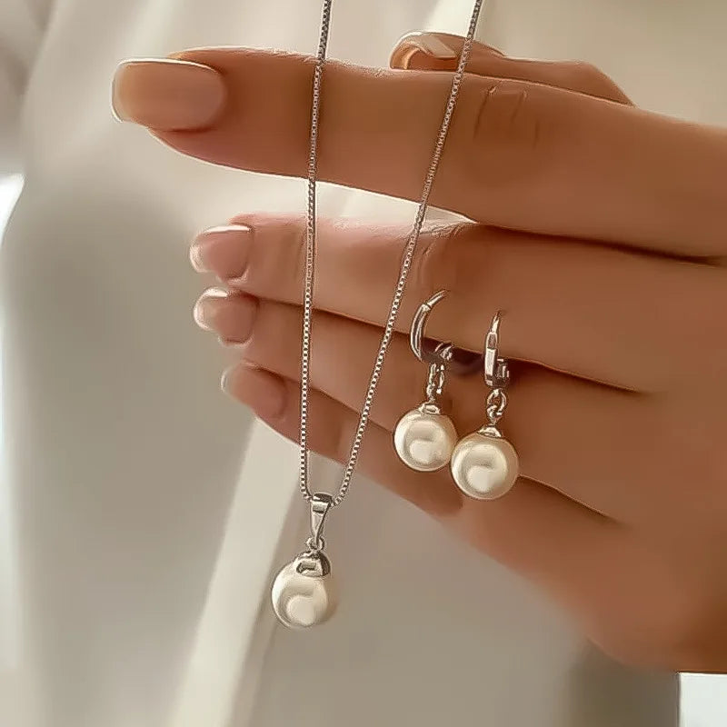 A hand models a dainty pearl jewelry set featuring matching earrings and a pendant necklace.