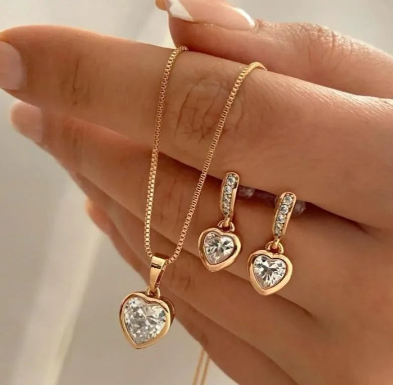 A hand displays a delicate gold necklace with a heart pendant and matching small heart-shaped earrings.