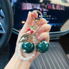 A hand holds a key chain decoration featuring a gold ring, leaves, and green and red pearl beads.