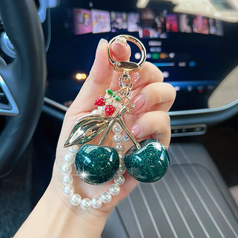 A hand holds a key chain decoration featuring a gold ring, leaves, and green and red pearl beads.